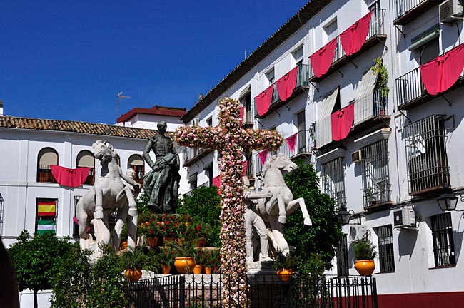 Maikreuz an der Plaza Conde de Priego am Denkmal des berühmten Stierkämpfers Manolete und blutroten Capas vor den Balkonen. Fiesta Cruzes de Córdoba, Andalusien, Spanien